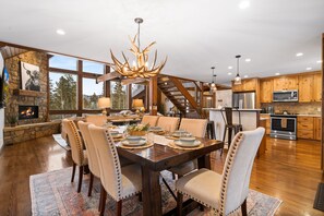 A spacious dining area with a wooden table, beige upholstered chairs, and a chandelier. A stone fireplace and large windows provide natural light.