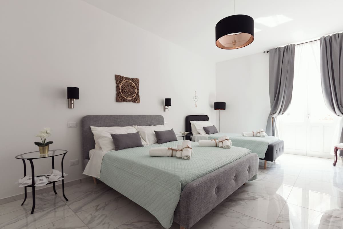 Et moderne hotelværelse med to senge, et rundt bord med en vase, og et stort vindue med gardiner.