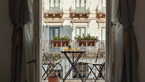 An open balcony with a table and chairs, potted plants, and a view of a building with balconies and windows. - Calafatari B&B (Syracuse)