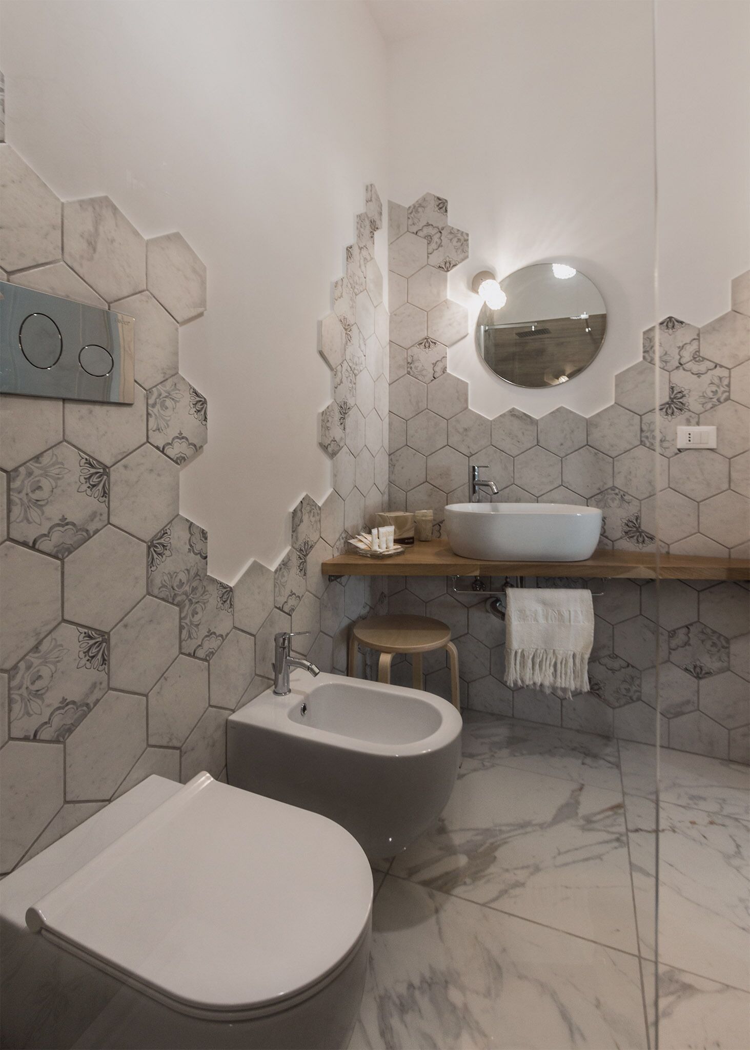 A bathroom with a hexagonal tile pattern, a round mirror, a sink, a bidet, and a toilet.