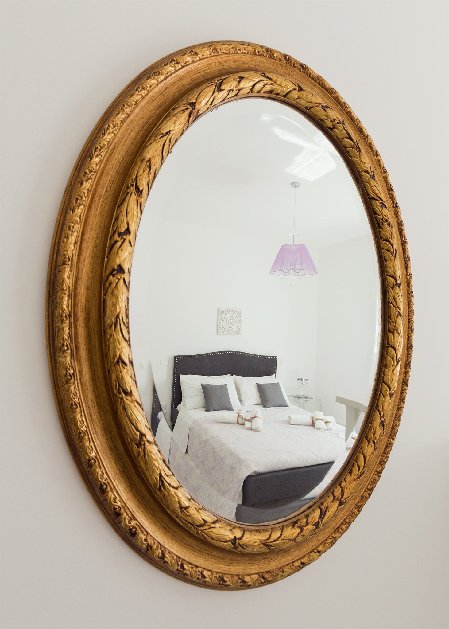 A round mirror with an ornate golden frame reflecting a bedroom with a bed, pillows, and a hanging light fixture.