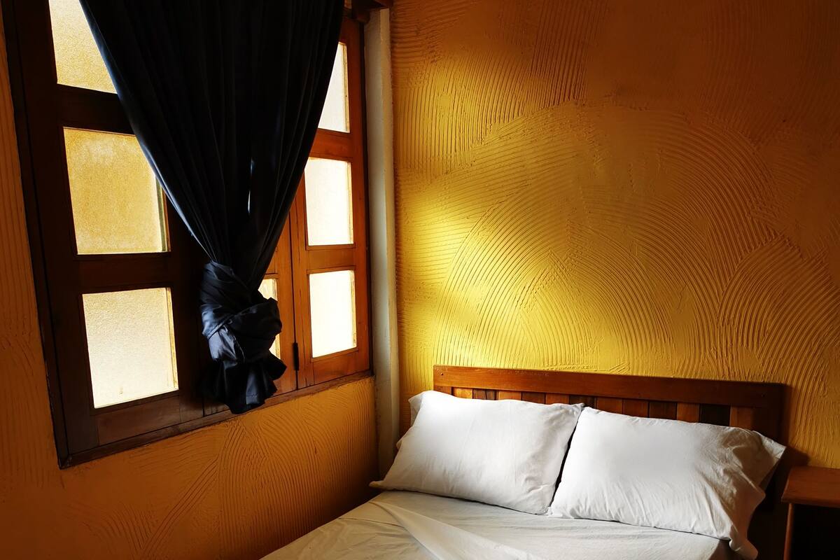 A single bed with white linens and a rolled towel, a wooden headboard, a nightstand, and a window with curtains.