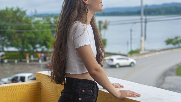 A person standing on a balcony overlooking a body of water with a road and vehicles in the background. - NATZ HOTEL (Flores)