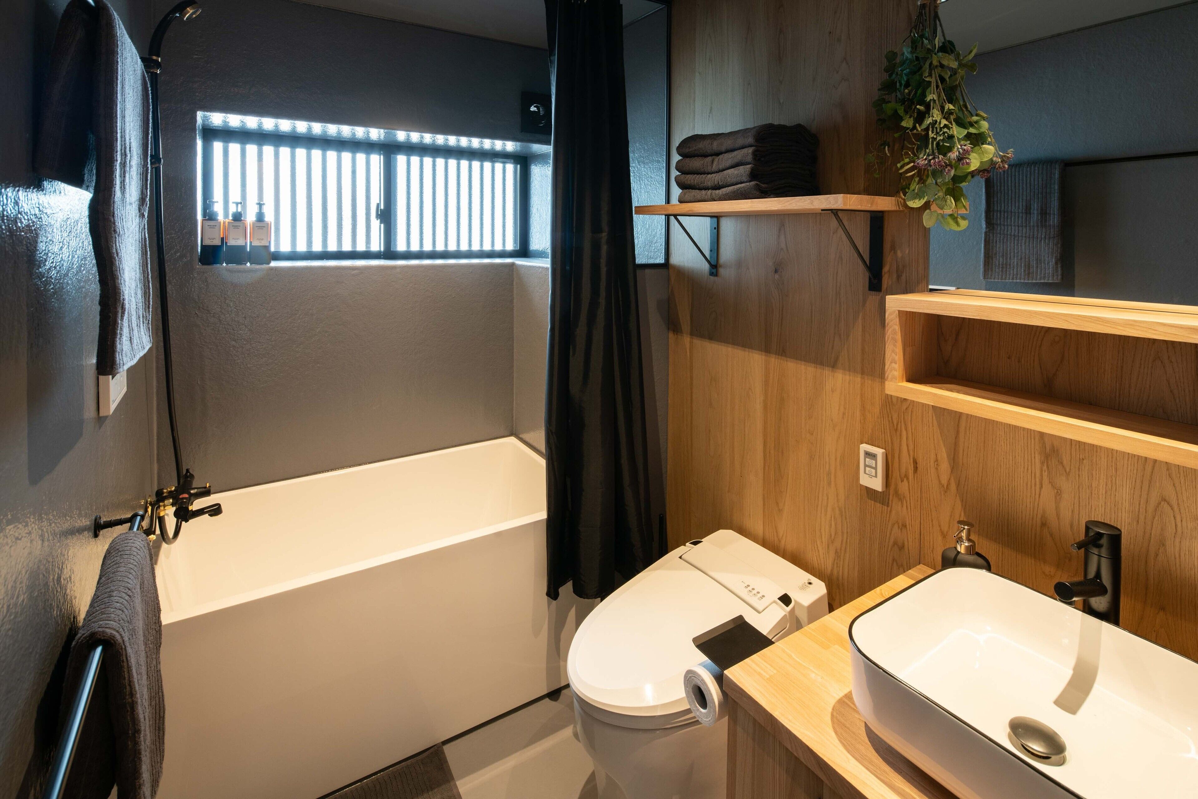 Un bagno moderno con una vasca, un water e un lavandino. Si vede una finestra con persiane, un porta asciugamani e uno scaffale con articoli da toilette.