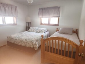 A bedroom with two beds, a window with a curtain, a bedside table with a lamp, and a door.