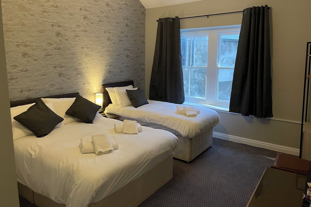 Quarto de hotel com duas camas, janela com cortinas, cabeceira e mesa de cabeceira.