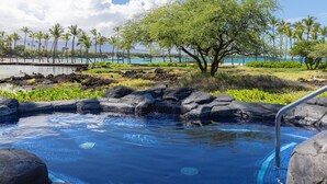 Ein Poolbereich mit klarem, blauem Wasser, umgeben von Felsen und einem Baum, mit Blick auf das Meer und Palmen im Hintergrund.