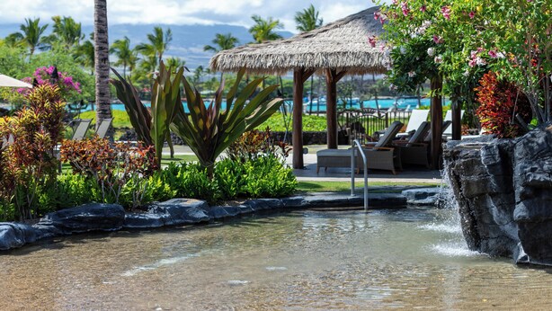 Un espace piscine de complexe hôtelier tropical, doté d’une structure à toit de chaume, de chaises longues et d’une petite cascade.