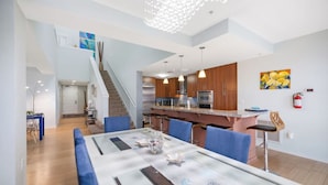 Una cocina moderna con electrodomésticos de acero inoxidable, un comedor con sillas azules y una escalera que conduce a un nivel superior.