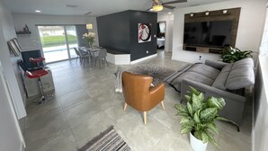 A modern living room with a grey sofa, a brown armchair, and a dining area with a glass table and chairs. - Modern lakeside pool home near hardrock and fll airport  (Davie)