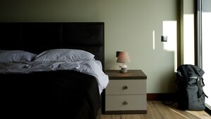 Uma cama bem arrumada com lençóis brancos, uma luminária de cabeceira, uma pequena mesa de cabeceira de madeira com gavetas e uma mochila preta no chão.