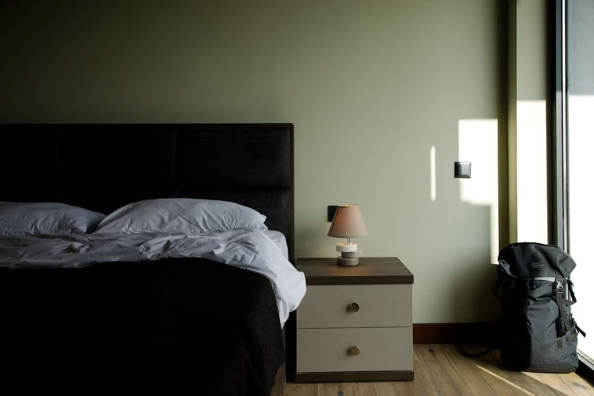 Una cama bien hecha con sábanas blancas, una lámpara de noche, una mesita de noche de madera con cajones y una mochila negra en el suelo.