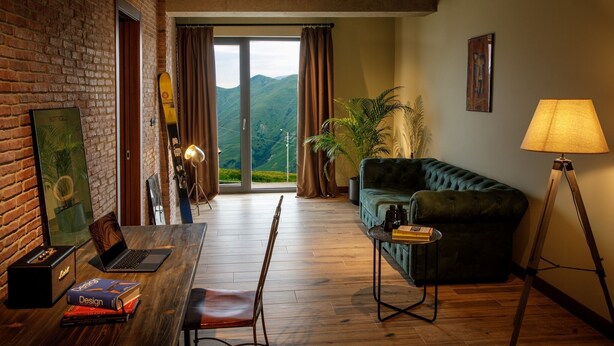 Quarto com piso de madeira, sofá verde, escrivaninha com laptop e livros, luminária e vista para montanhas por uma porta de vidro.