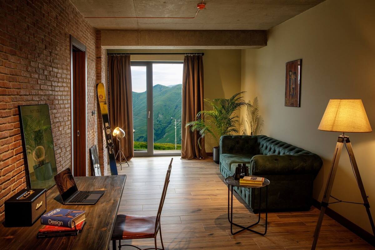 Una habitación con piso de madera, un sofá verde, un escritorio con una computadora portátil y libros, una lámpara y vistas a montañas a través de un ventanal.