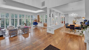 A spacious living room with hardwood flooring, white walls, and large windows. There is a dining area with a wooden table and chairs, and a seating area with sofas and chairs. - Fancy Poconos Castle & Indoor MEGA 10+ Hot Tub (East Stroudsburg)