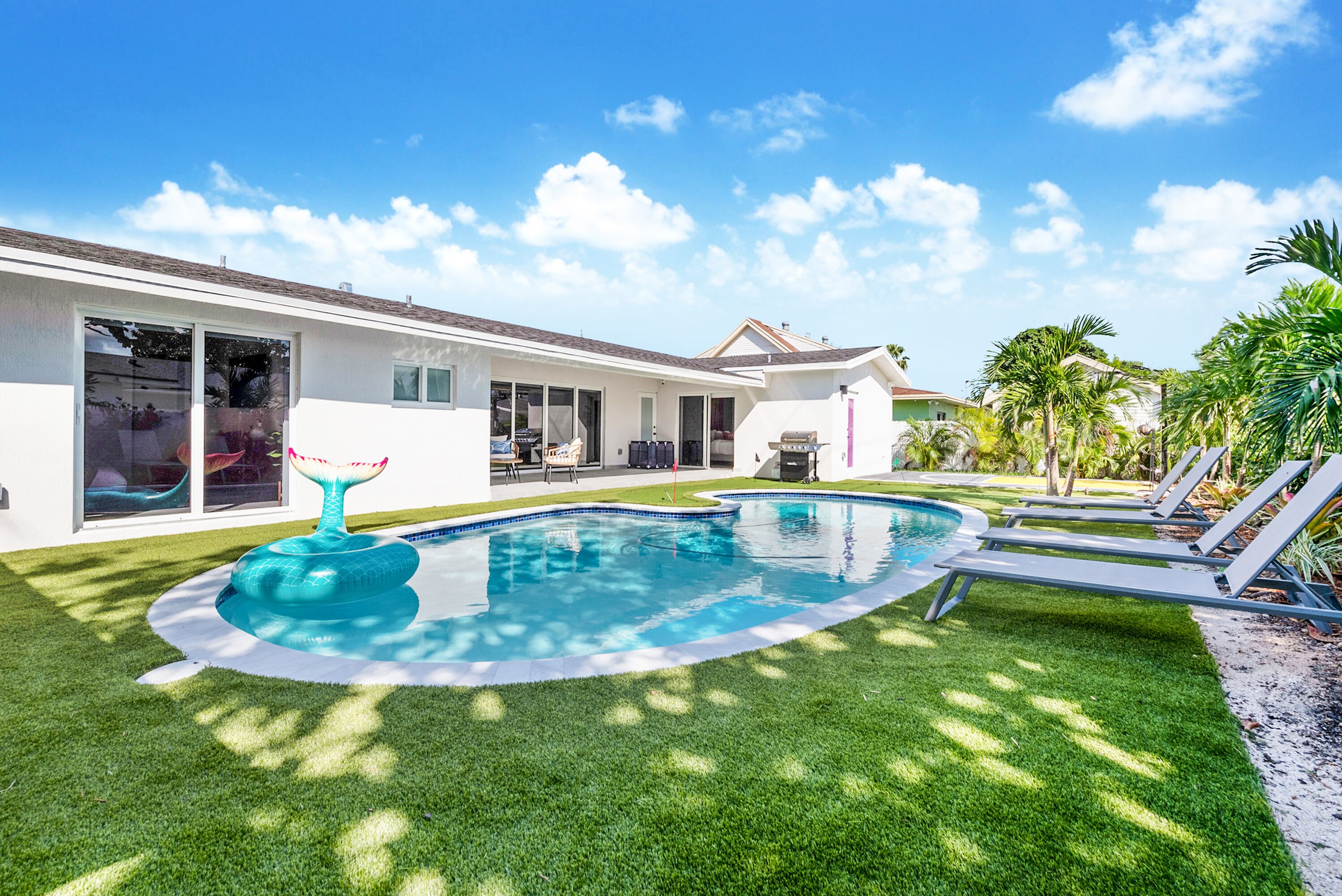A backyard with a swimming pool, a lawn, and a house with large windows and a patio.