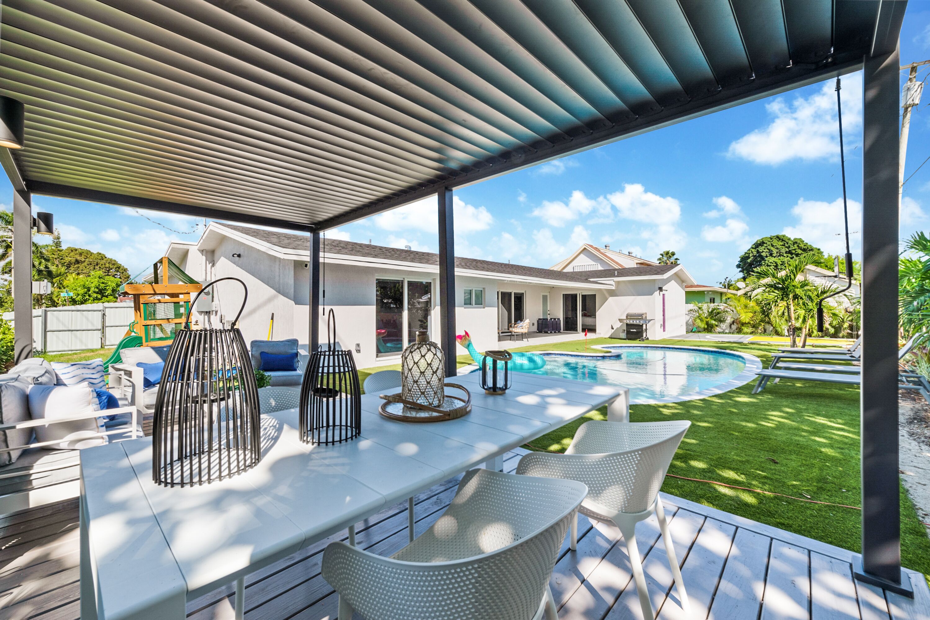 Et overdækket udendørs område med en pool, hvide fletmøbler og et pergola.