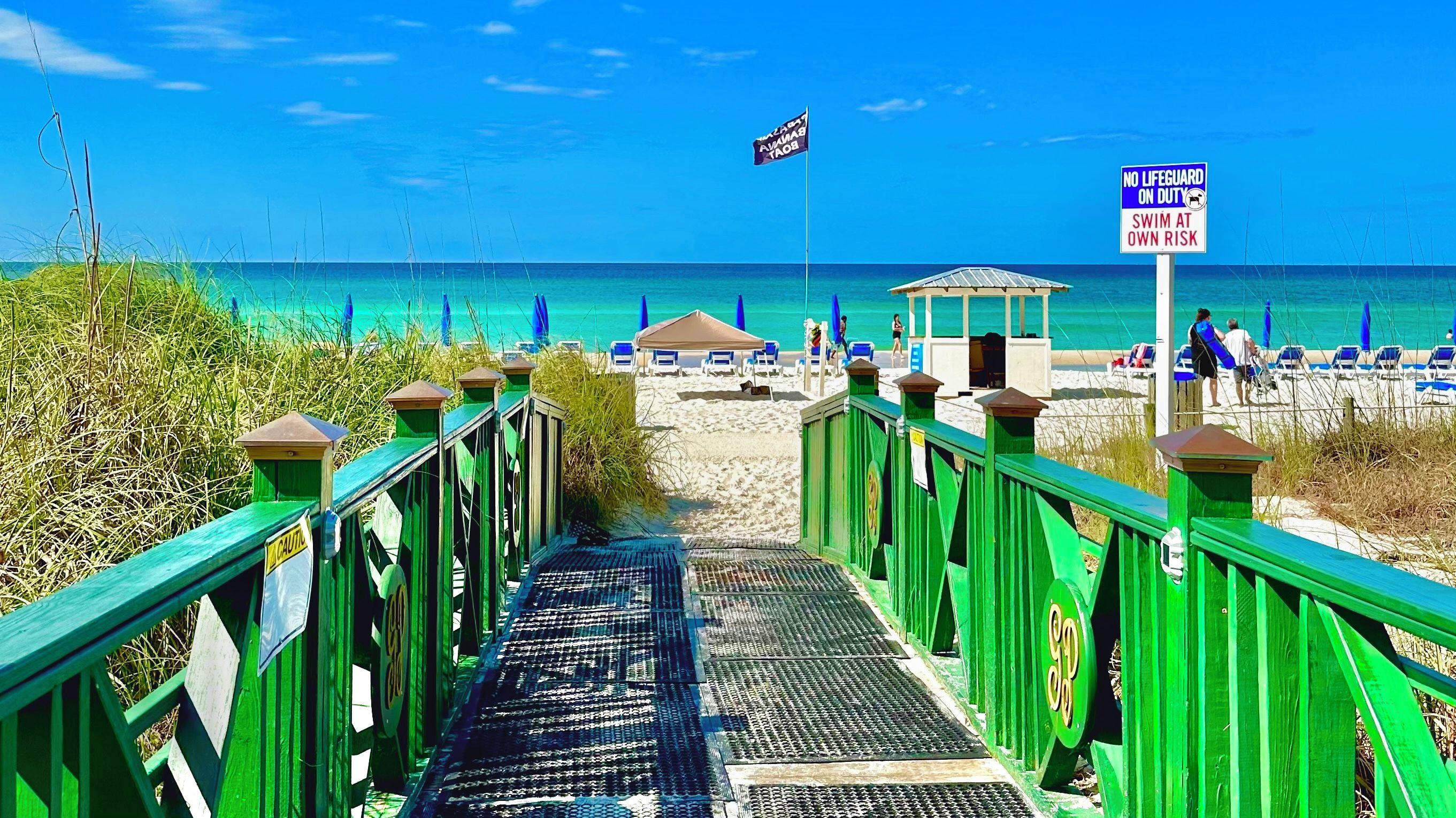 Ein Gehweg führt zu einem Sandstrand mit Sonnenschirmen und Liegestühlen.