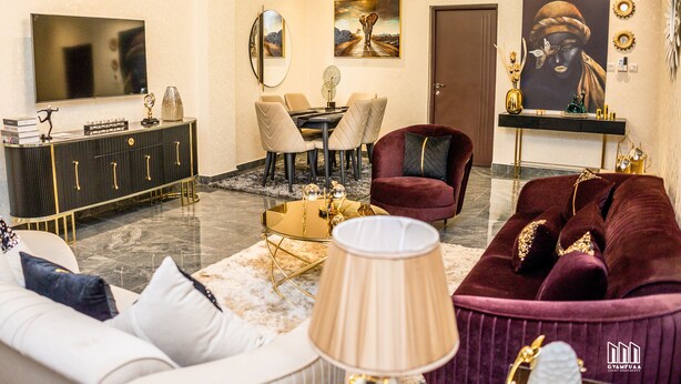 A modern living room with a sofa, armchair, and a coffee table. There is a TV mounted on the wall, a dining table with chairs, and a door. - Gyamfuaa Court Apartment (Kumasi)