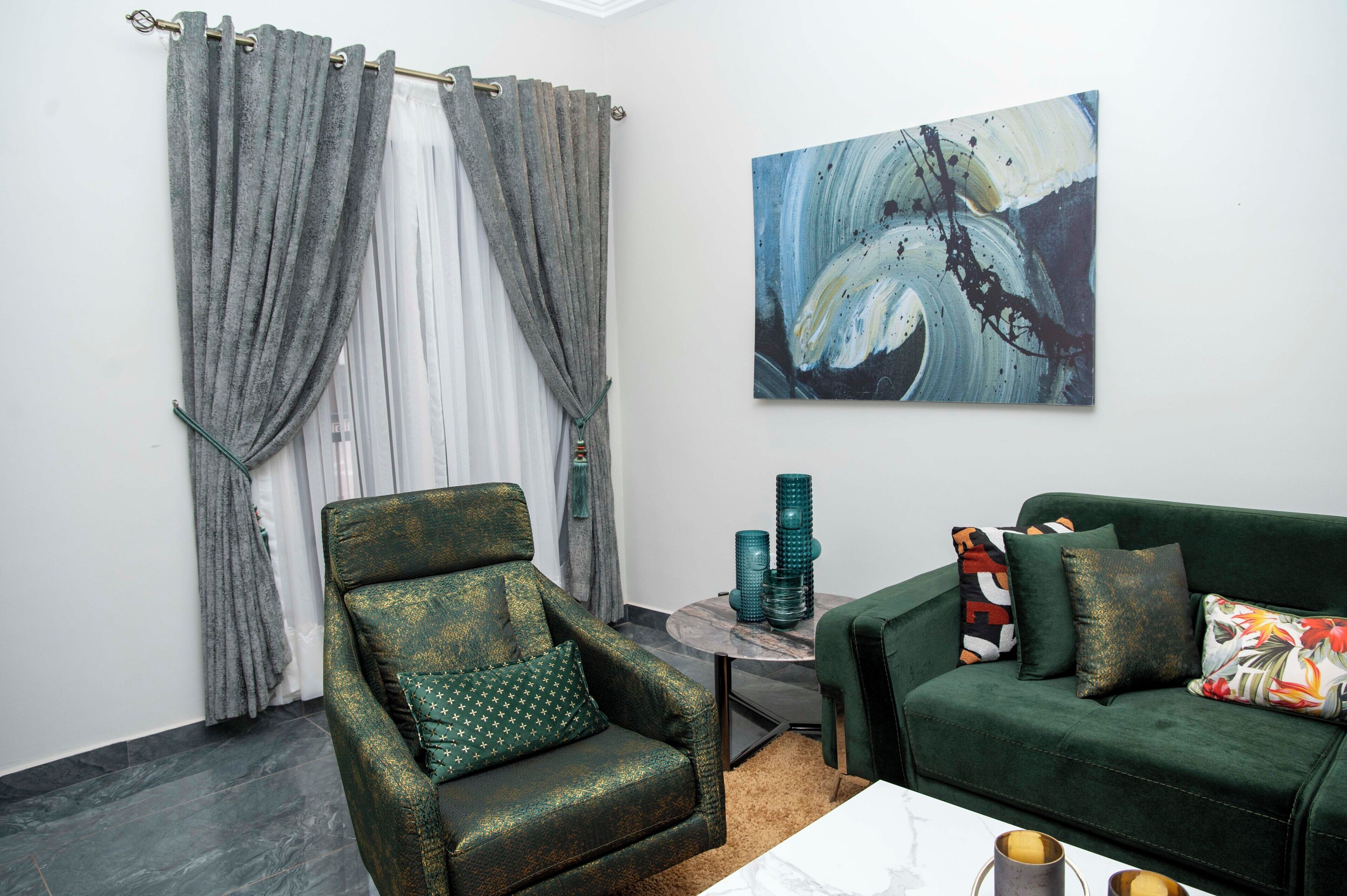 A room with a green armchair, a green sofa, a round side table with decorative items, and a painting on the wall.