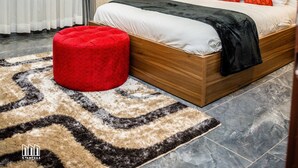 A modern bedroom with a wooden bed, red and orange pillows, a red ottoman, a painting on the wall, and a patterned rug. - Gyamfuaa Court Apartment (Kumasi)