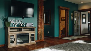 A modern living room with a large flat-screen TV mounted on the wall, wooden flooring, and a large rug. - Connoisseur Hideaway (Bozeman)