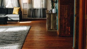 A spacious living room with a wooden floor, a sofa, a coffee table, and a rug. There are two windows with sheer curtains, a floor lamp, and a wooden door. - Connoisseur Hideaway (Bozeman)