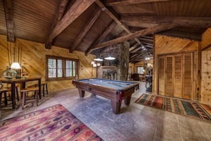 Interior de madeira com mesa de bilhar, área de refeições e lareira de pedra.