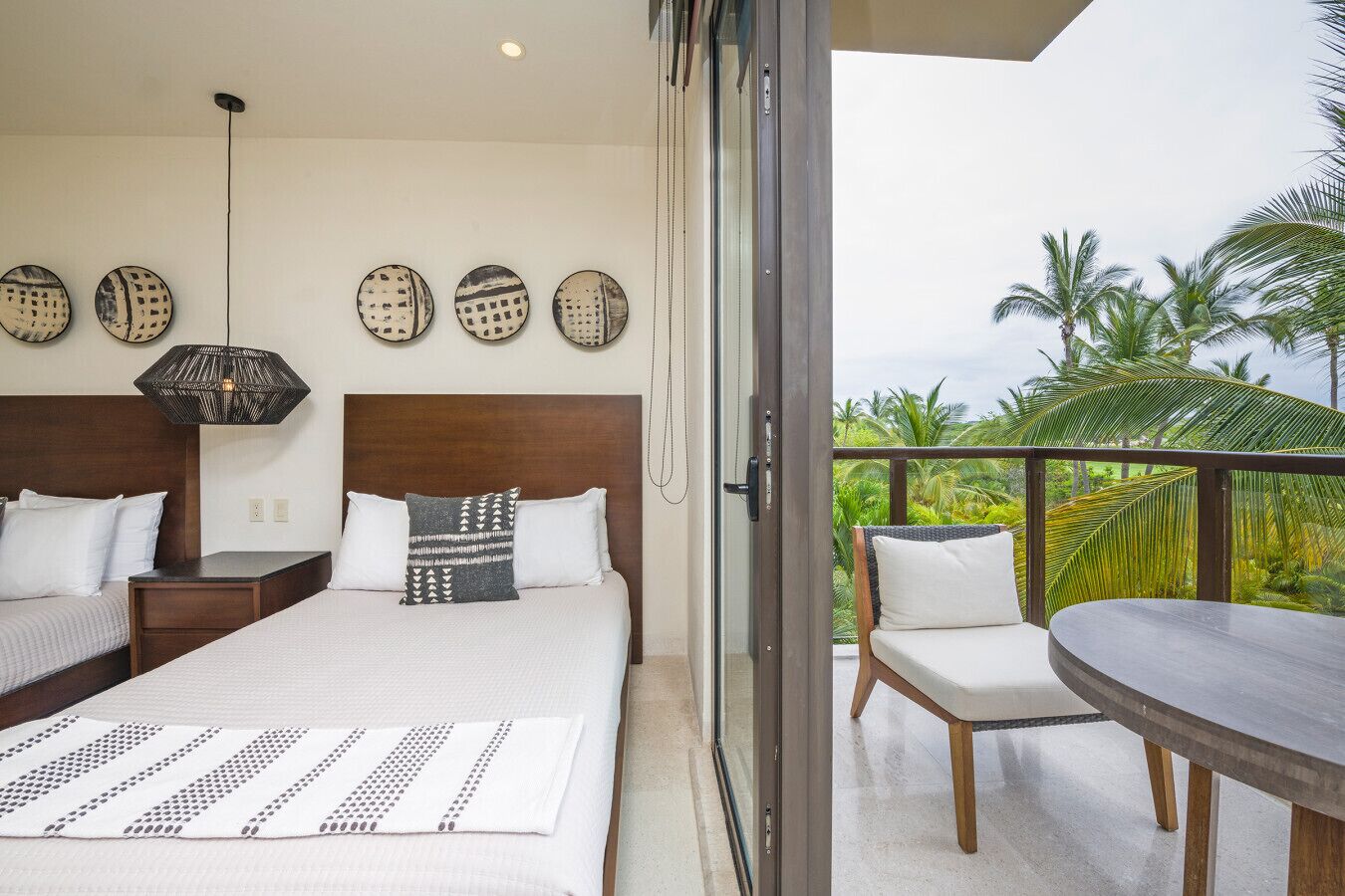 A bedroom with a large bed, a nightstand, a bedside table, and a chair. There is a balcony with a table and chairs, and a view of palm trees.