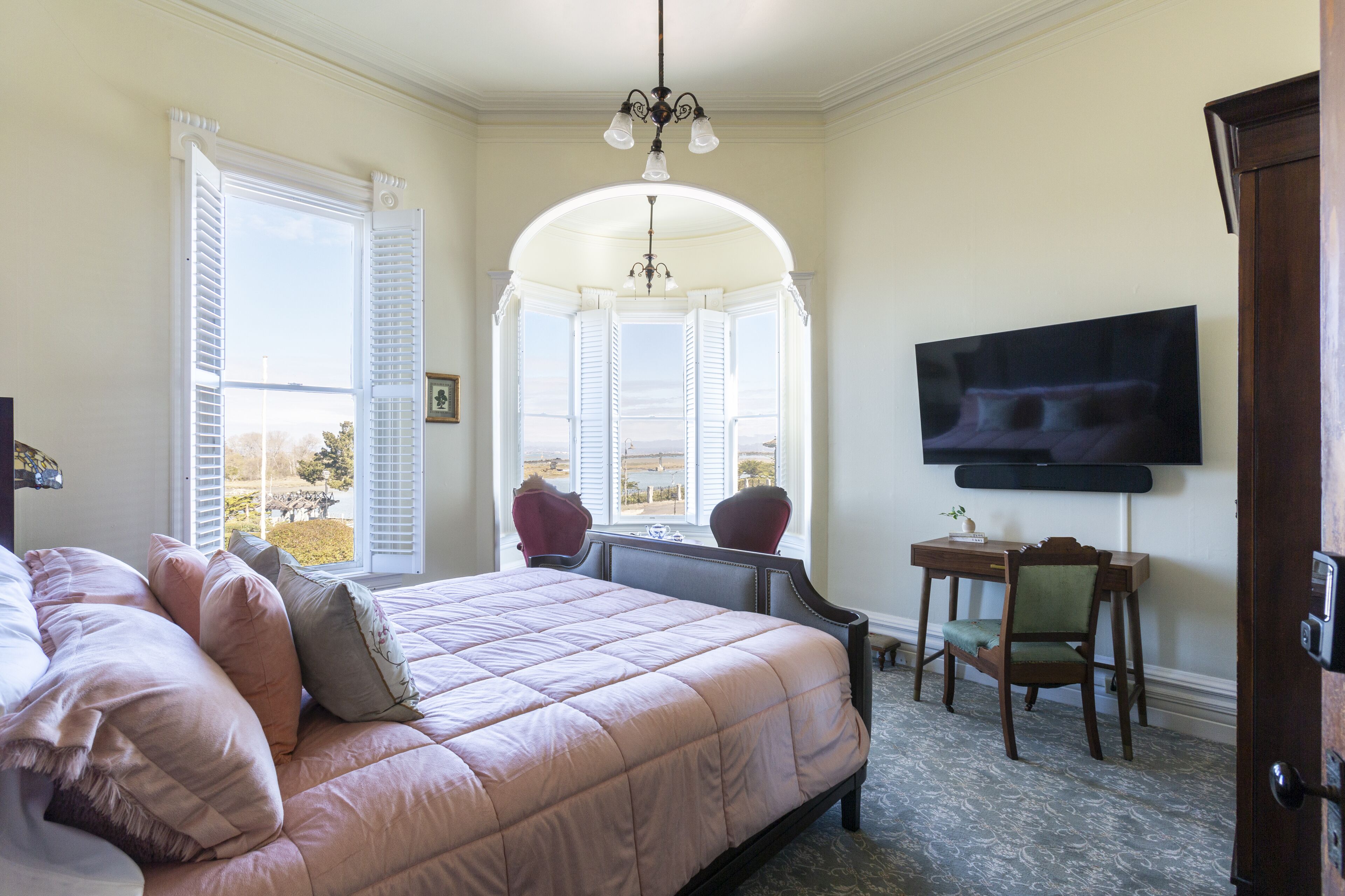 A bedroom with a large bed, a bay window, a television, and a desk.
