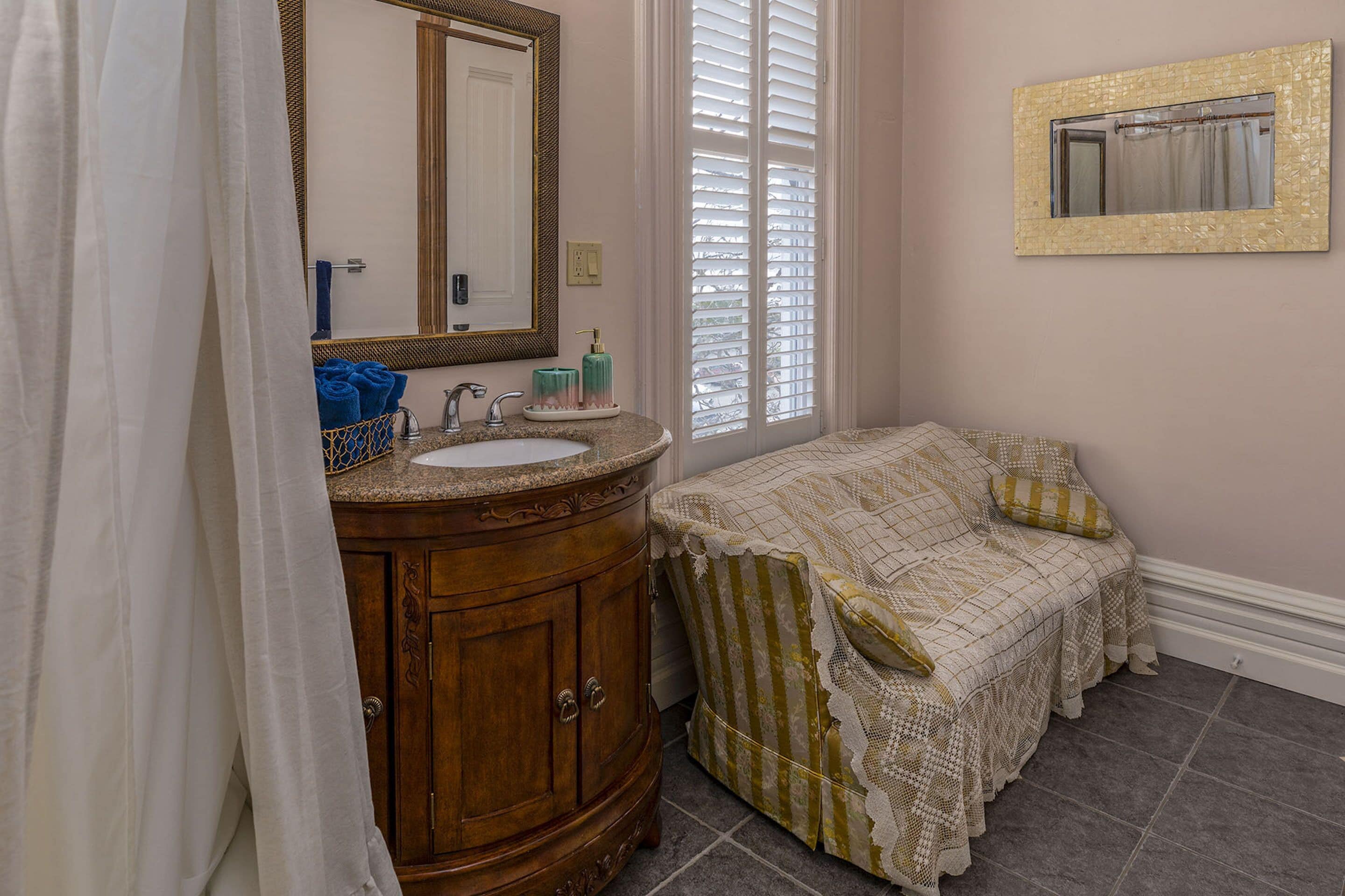 A bathroom with a wooden vanity, a large mirror, a window with blinds, and a bed with a patterned bedspread.