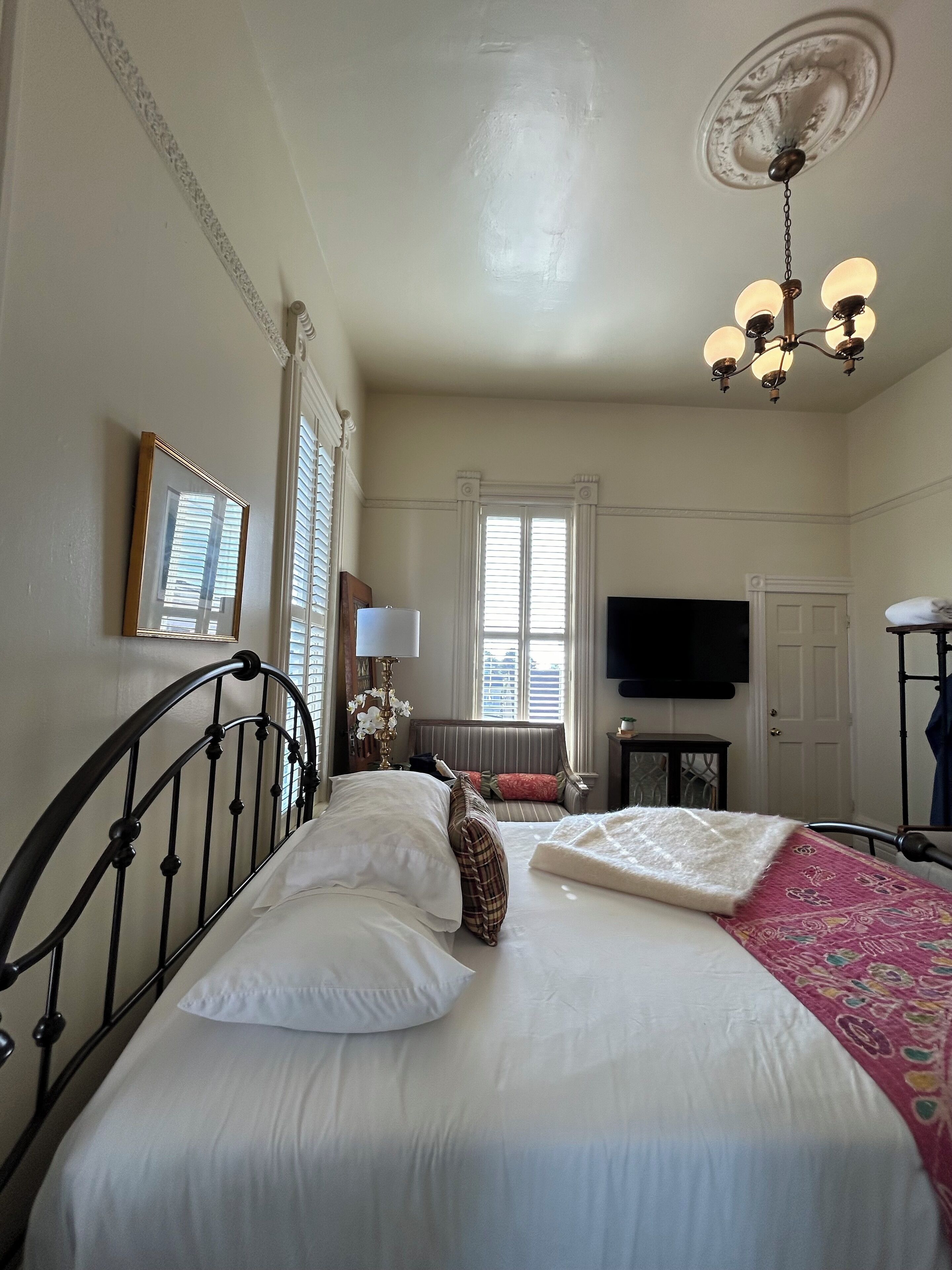 Una habitación de hotel con cama, televisor, candelabro y ventana con persianas.