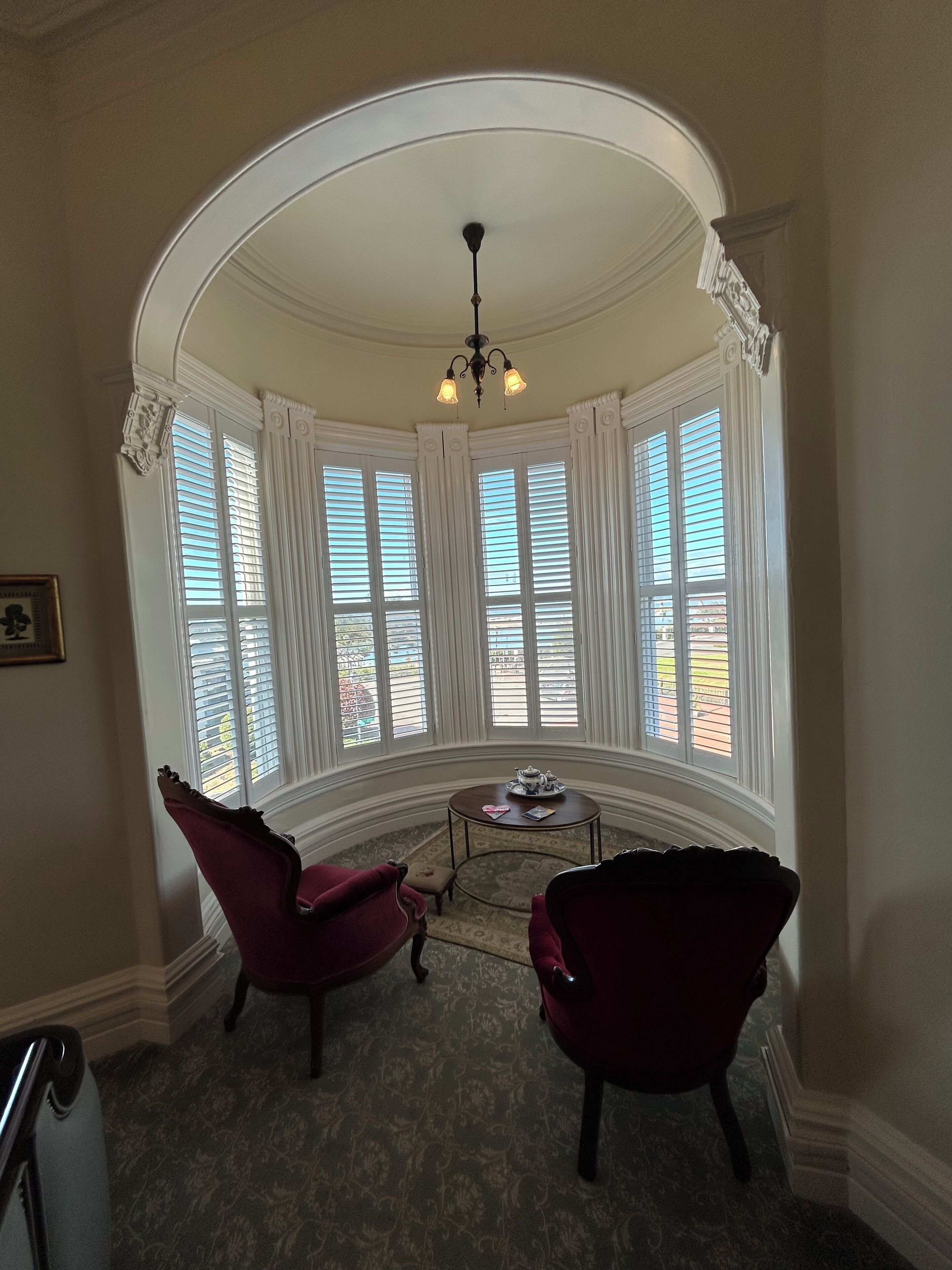 A room with a curved ceiling, large windows with shutters, a chandelier, two red armchairs, a small table with a teapot, and a framed picture on the wall.