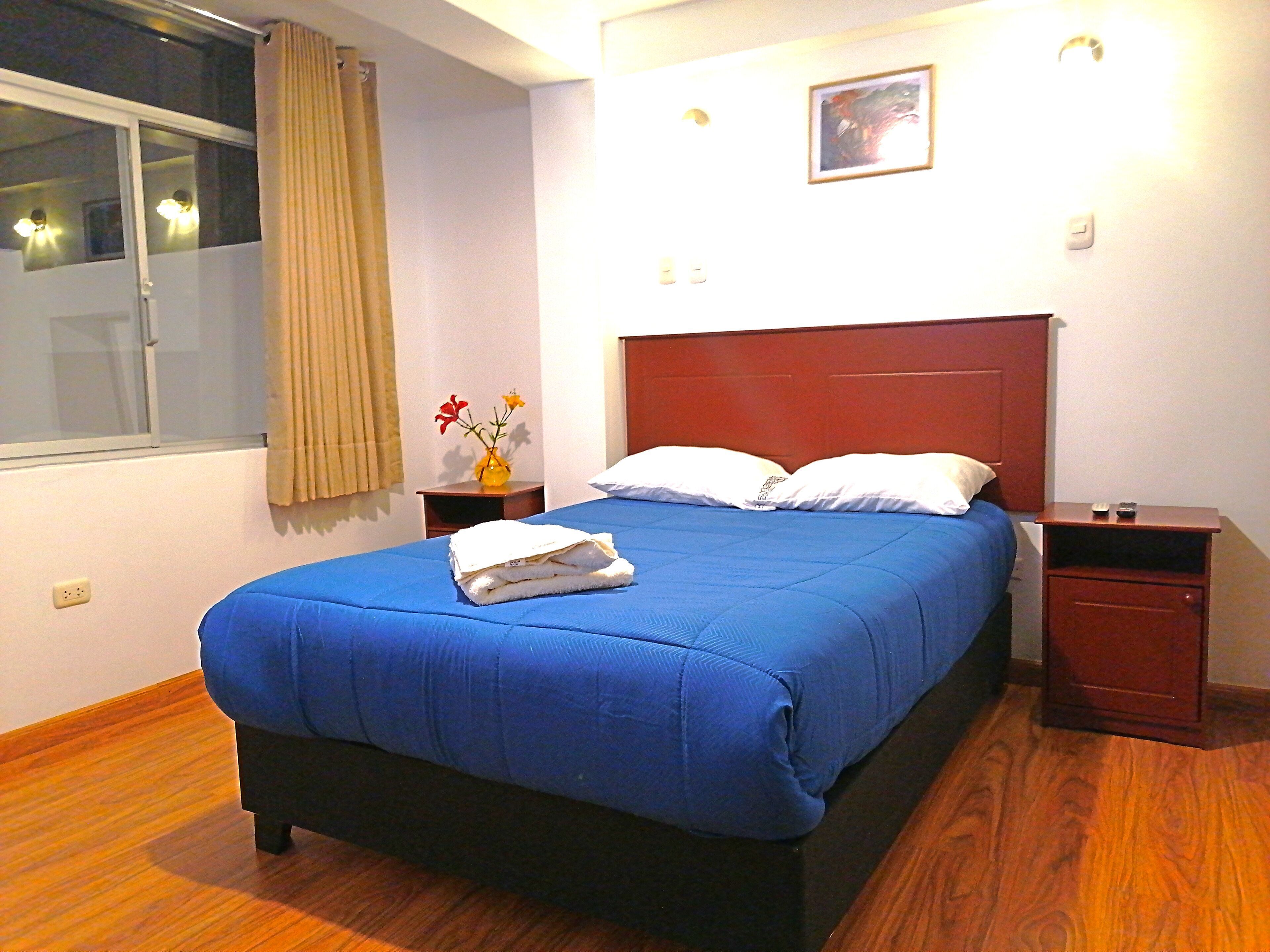 A neatly made bed with a blue bedspread, a vase with flowers, and a nightstand with a remote control.