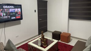 Un salon équipé d’un téléviseur à écran plat affichant l’interface d’un service de diffusion en continu, d’une table basse en verre, d’une porte et d’une fenêtre avec des stores.