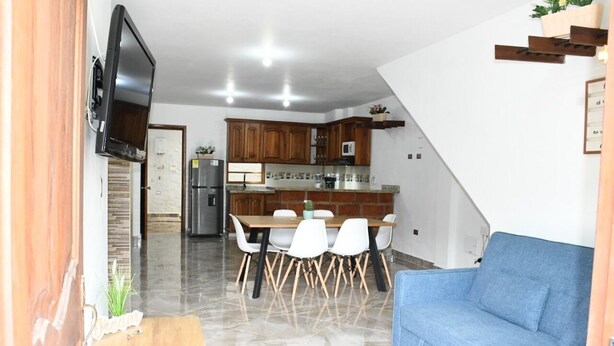 Uma cozinha moderna com armários de madeira, uma mesa de jantar com cadeiras brancas, um sofá azul e uma TV fixada na parede.