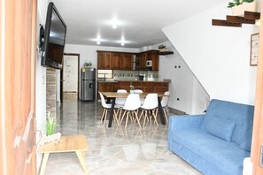 Eine moderne Küche mit Holzschränken, einem Esstisch mit weißen Stühlen, einem blauen Sofa und einem an der Wand befestigten Fernseher.