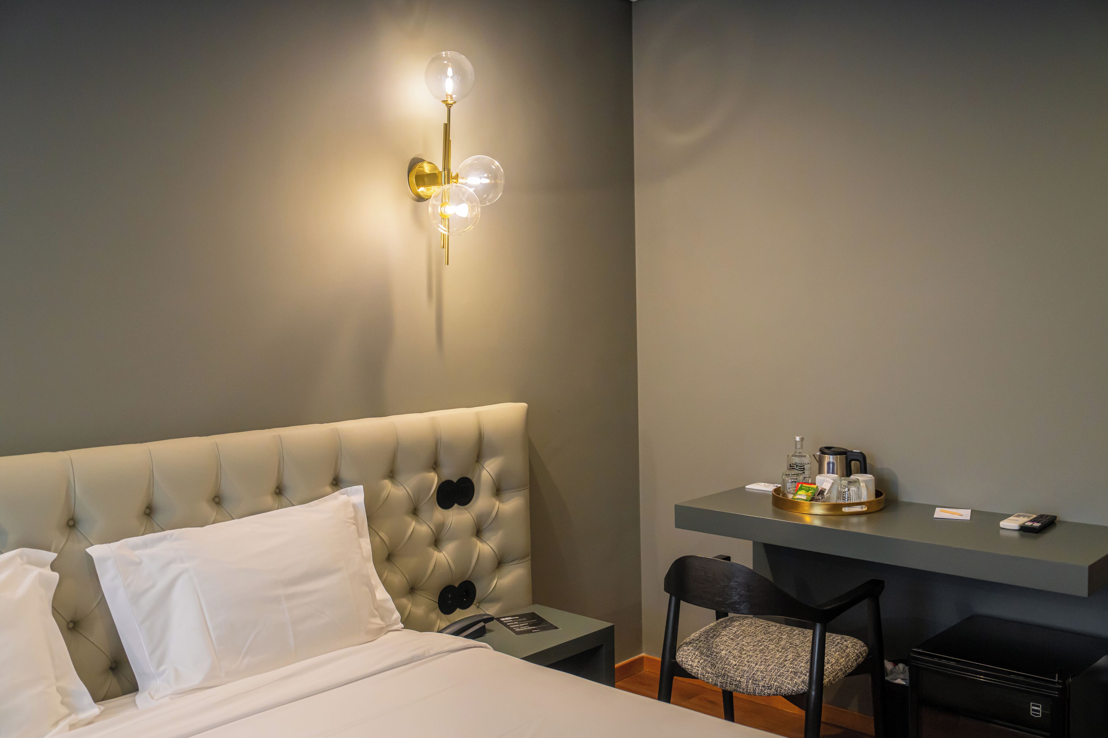Una camera d'albergo con una testata imbottita, un comodino con un vassoio di articoli da toilette, una scrivania con sedia e un lampadario a parete.