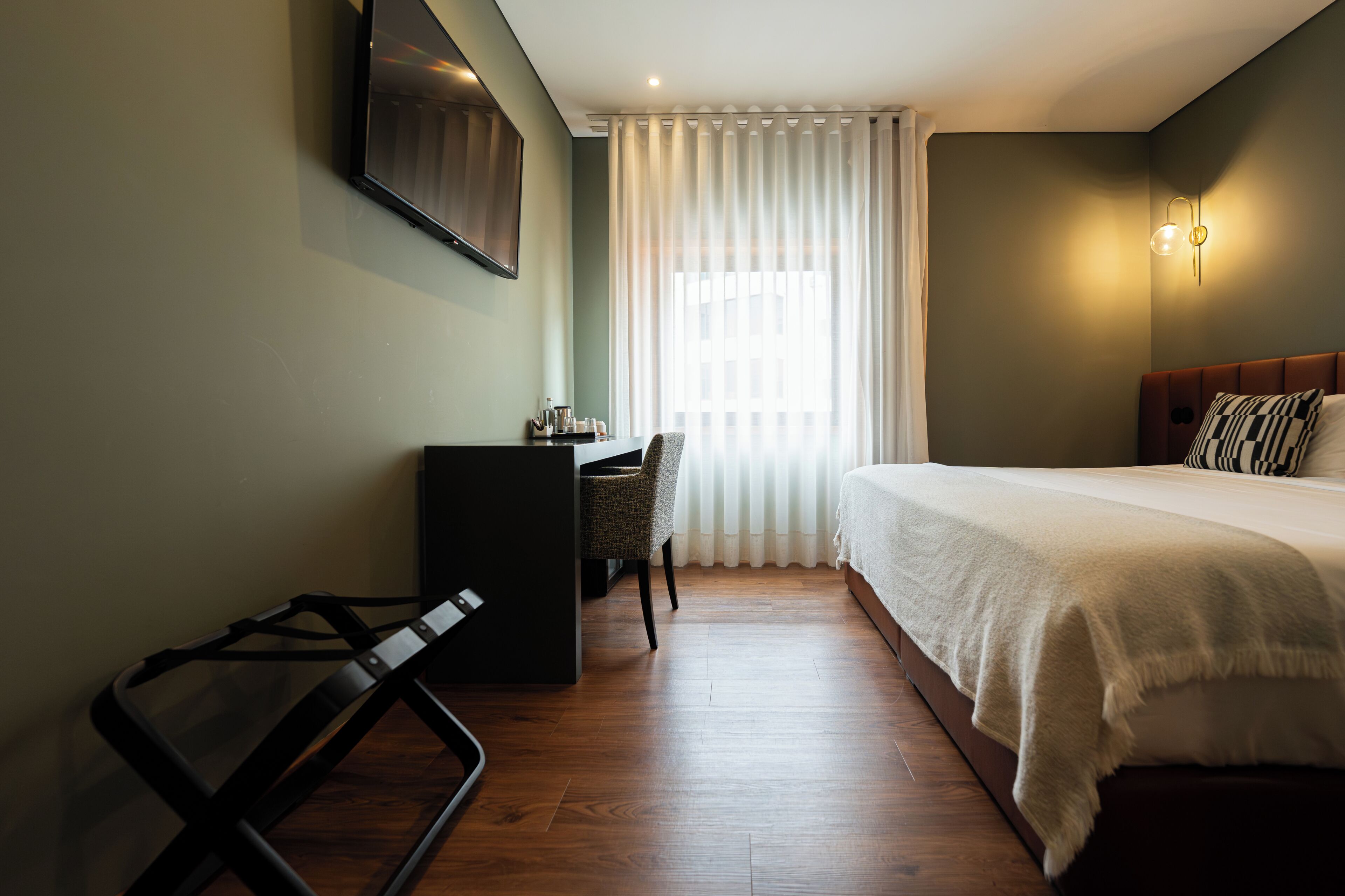Une chambre d’hôtel comprenant un lit, un bureau, une chaise, un téléviseur fixé au mur et une fenêtre avec des rideaux.