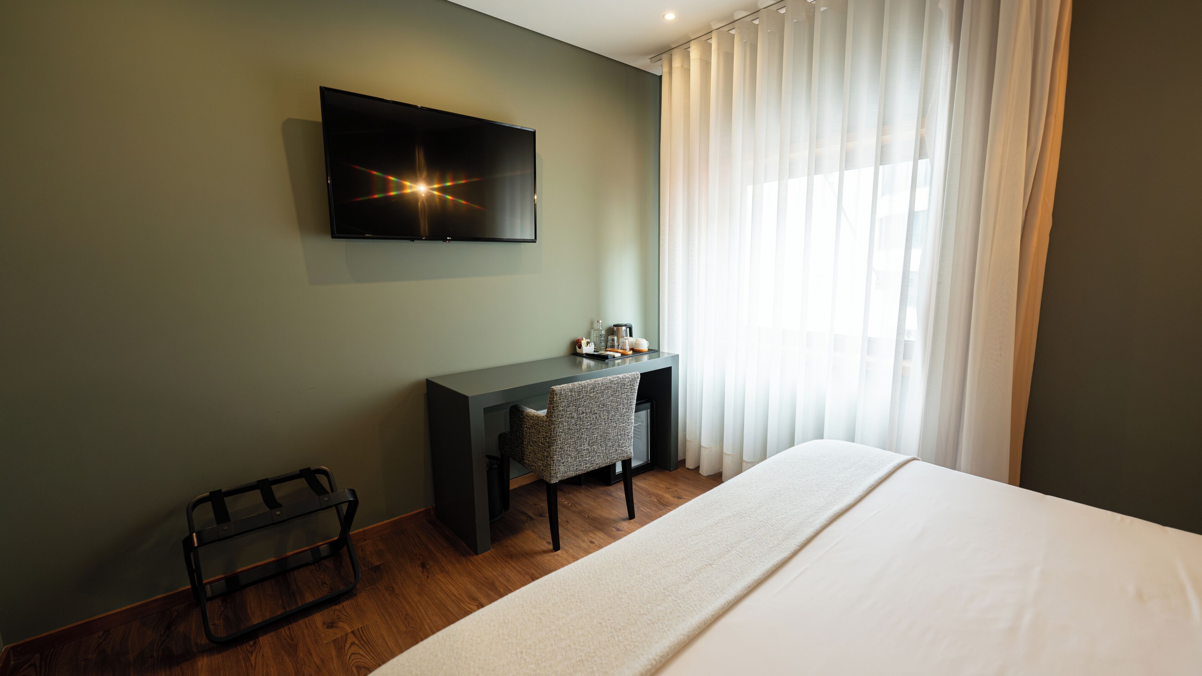 Habitación de hotel con cama, escritorio, silla, televisor montado en la pared y ventana con cortinas.