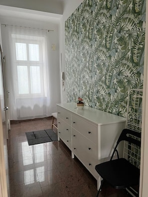 Une pièce avec une commode blanche, une chaise noire, une fenêtre avec des rideaux blancs et un mur orné d’un motif de feuilles tropicales.