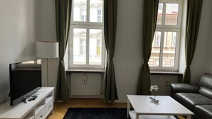 Ein modernes Wohnzimmer mit Fernseher, einem weißen Couchtisch, einem Ledersofa und zwei Fenstern mit Vorhängen.