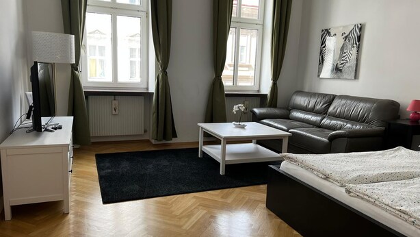 Ein modernes Wohnzimmer mit einer Couch, einem Couchtisch, einem Fernseher auf einem Lowboard und einem Bett mit weißer Bettdecke.