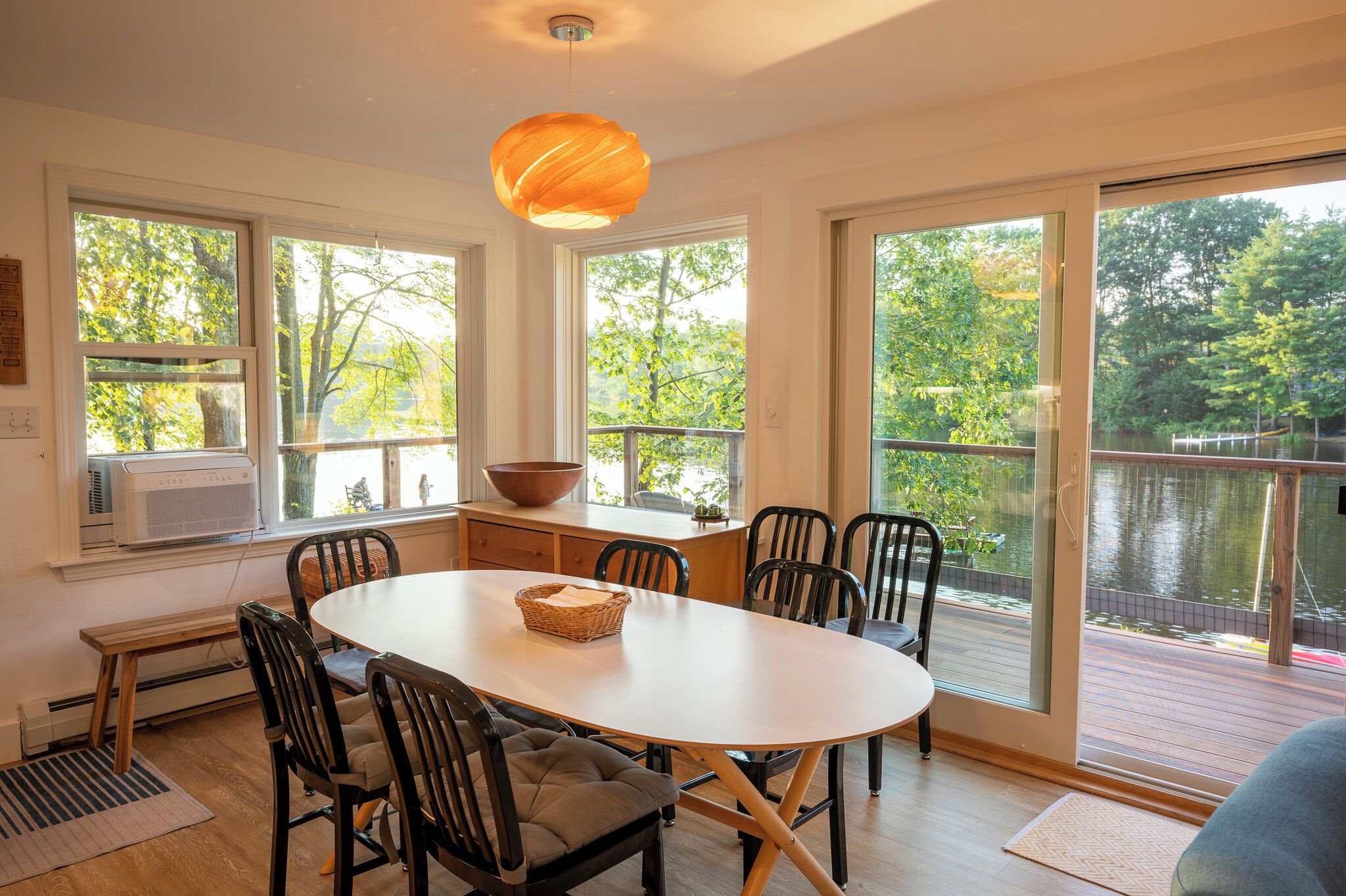Un coin repas avec une table ronde, des chaises, un luminaire suspendu et de grandes fenêtres donnant sur un lac et des arbres.