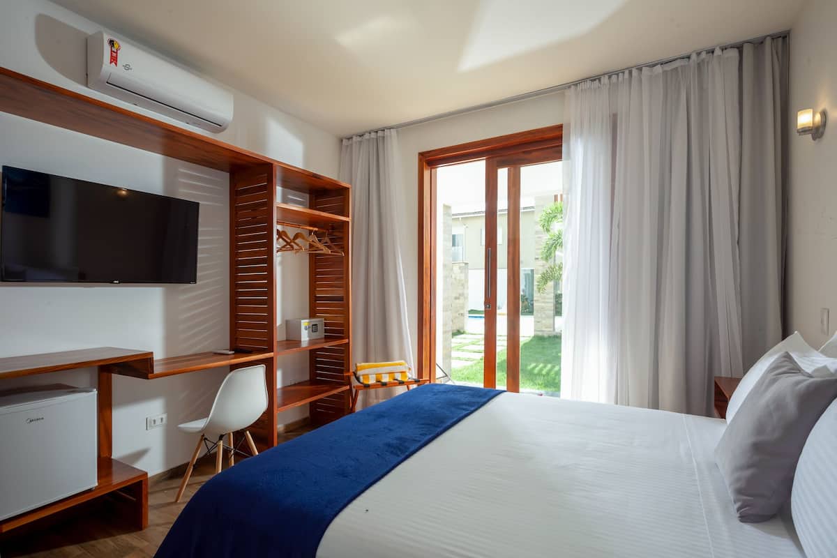 Quarto de hotel com cama, televisão, escrivaninha, guarda-roupa e vista para um jardim.