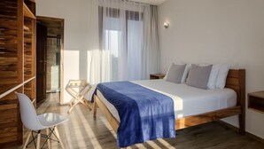 A bedroom with a wooden bed, a white chair, a small table, and a window with sheer curtains. - AIOLOS HOTEL & DISCOVERIES (Cruz)