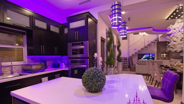 A modern kitchen with dark cabinetry, a central island, and a dining area with purple chairs. - 🕯Versaille’s Residence ™ (McAllen)