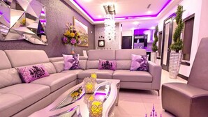 A modern living room with a sectional sofa, a glass coffee table with decorative items, and a chandelier. - 🕯Versaille’s Residence ™ (McAllen)