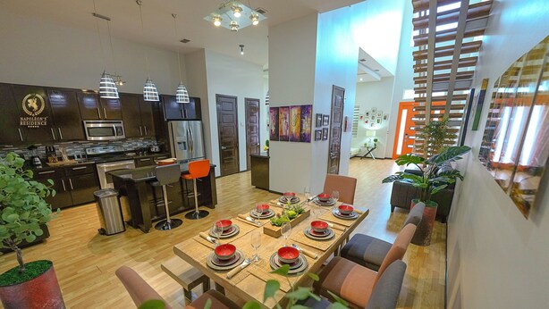 Cocina moderna con armarios oscuros, un microondas y un refrigerador de acero inoxidable. Hay una zona de comedor con una mesa de madera dispuesta para seis personas, rodeada de sillas. El ambiente tiene un ventanal y vistas al exterior.