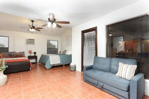 Un salon bien éclairé, avec un canapé, un lit recouvert d’une couverture verte, un ventilateur de plafond et de grandes fenêtres équipées de stores.